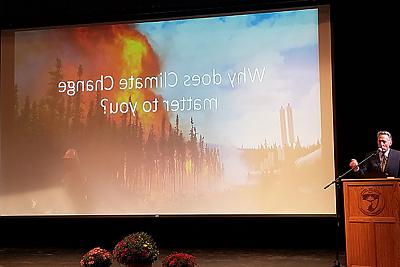 前佛蒙特州州长彼得·舒姆林站在格林霍剧院的讲台上. 屏幕投影显示了森林火灾的图像和“为什么气候变化对你很重要”的文字? 
